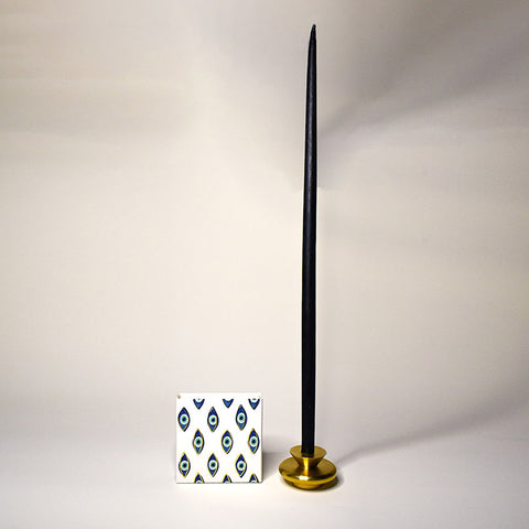 A brass candle holder holding a tall black candlestick with a pack of matches in evil eye packaging next to it.