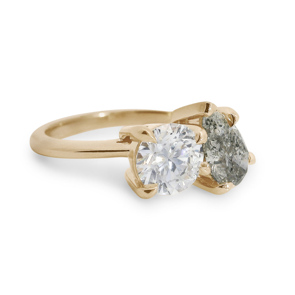 Two diamond toi et moi ring featuring a round brilliant white diamond and a pear shaped grey diamond with gold setting on a white background