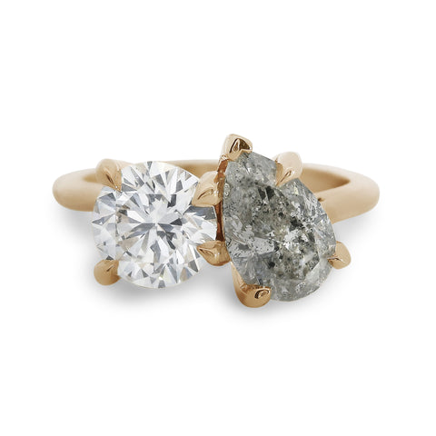 Two diamond toi et moi ring featuring a round brilliant white diamond and a pear shaped grey diamond with gold setting on a white background