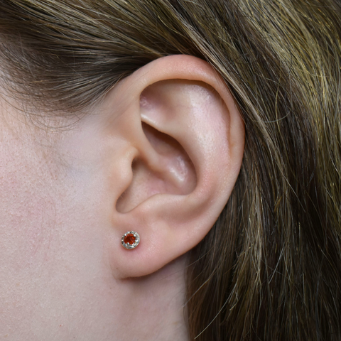 Close-up of an ear wearing a small round earring with a red-orange gemstone. 2