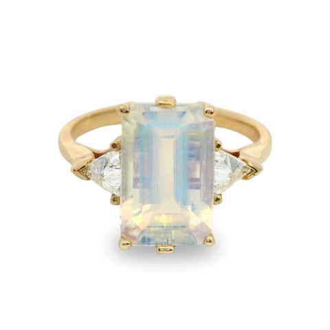 A three stone yellow gold ring featuring an emerald cut rainbow moonstone center and two white diamond trillion side stones on a white background