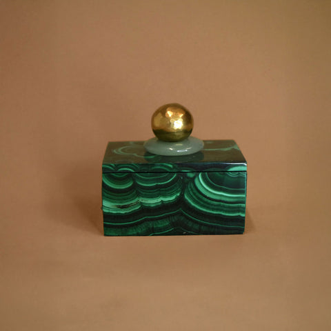 Small malachite box with an Aventurine gemstone and bronze ball on top of lid on a beige background