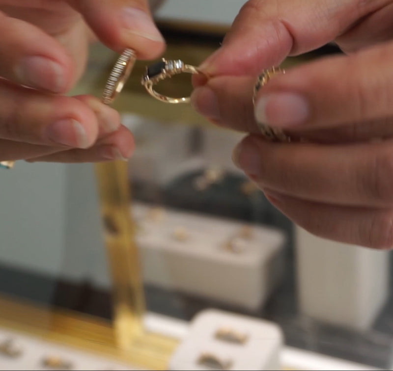 Zoomed in view of two hands holding a five stone black diamond ring and a straight white diamond band over a jewelry case.