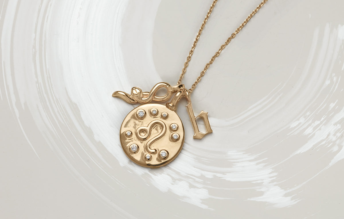 alt="A bird's eye view of a Pendant with a Leo symbol surrounded by white diamonds, paired with a letter "b" charm and serpent charm on a chain."