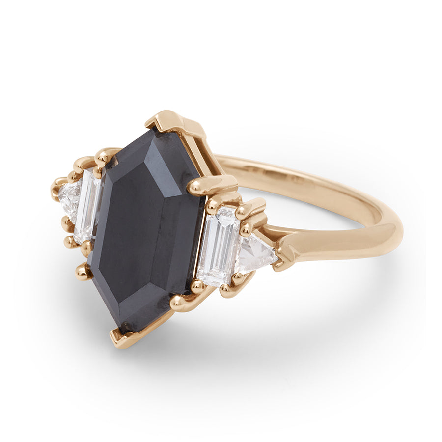 Angled view of a ring with a hexagonal shaped black diamond center stone with one baguette white diamond and one white diamond trillion on either side set in yellow gold.