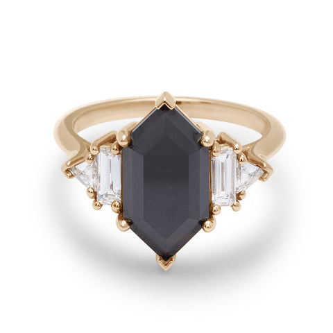 A ring with a hexagonal shaped black diamond center stone with one baguette white diamond and one white diamond trillion on either side set in yellow gold.