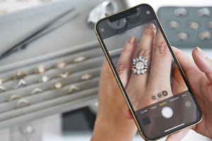 A medium complexion hand wearing a white diamond solitaire ring with two stacking bands holding an iPhone over two trays of assorted jewelry.