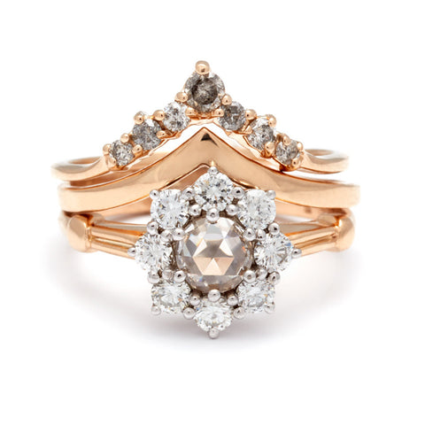 A cluster engagement ring with a rose cut champagne diamond center stone and round white diamond stones surrounding it set in yellow gold stacked beneath a grey diamond nesting band and a solid gold chevron band.