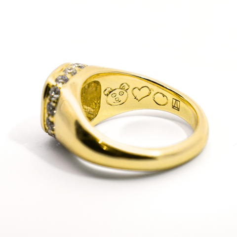 A side angle of a white diamond halo yellow gold signet ring showing specifically a custom engraving on the inside of the band that depicts the face of a teddy bear, a heart, and a circle next to an Anna Sheffield Jewelry stamp. 