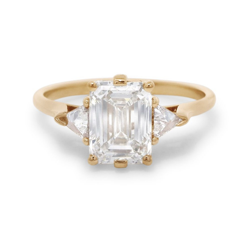 A yellow gold three stone engagement ring featuring an emerald cut diamond center and two trillion diamond side stones on a white background