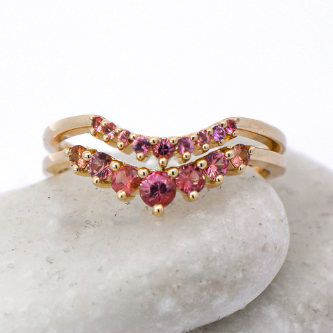 Two curved women's wedding bands featuring Padparadscha Sapphires set in yellow gold. The bands sit on a white stone against a white background. 