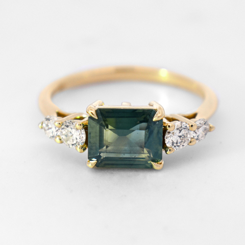 A five stone ring featuring an emerald cut green sapphire and two round diamond side stones on either side set in yellow gold. 
