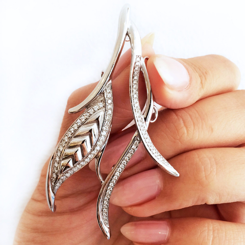 A hand with almond shape pink-nude nails holding a custom white gold wedding day hairpiece. The hairpiece  is split into two wispy strands that depict the silhouette of a leaf and encrusted with white diamonds. 