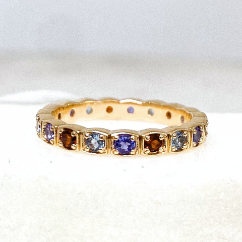 A gemstone eternity band featuring alternating red, blue, and purple gemstones set in yellow gold against a white background. 