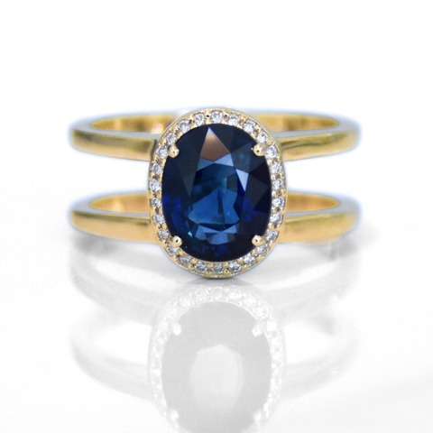 A ring featuring an oval cut blue sapphire encircled with a white diamond halo and set on double round yellow gold bands against a white background. 