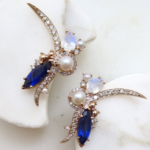 Two custom-made earrings in the organic shape and silhouette of lying flowers, featuring pearls, rainbow moonstone, and a large marquise shape blue gemstone and round white diamonds set in yellow gold. 