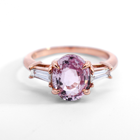 A three stone ring featuring a round pink morganite center stone and white tapered baguette diamond side stones set in rose gold against a white background. 