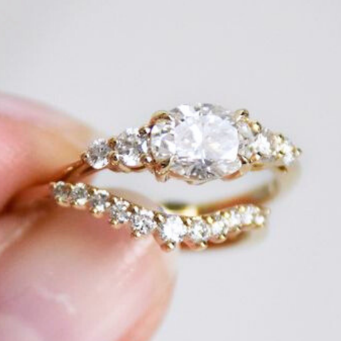 A photo of two rings between someone's index finger and thumb. The above ring is a five stone engagement ring featuring an East-West set white oval diamond paired with two white round diamonds on either side set in yellow gold. The bottom ring is a nesting band to stack perfectly with the top ring. This ring features graduated white round diamonds set in yellow gold on a slight curve. 