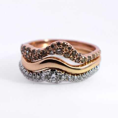 Three bands meant to be stacked and sit flush on the finger. The top is a curvy wave-shaped rose gold band with round grey diamonds set. The middle is a yellow gold band of the same shape. The bottom one is sitting flush with the above band but has less of an intense wave. It's a white gold band featuring round grey diamonds. 