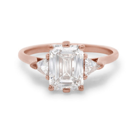 A rose gold three stone engagement ring featuring an emerald cut diamond center and two trillion diamond side stones on a white background 4