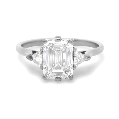 A white gold three stone engagement ring featuring an emerald cut diamond center and two trillion diamond side stones on a white background 3