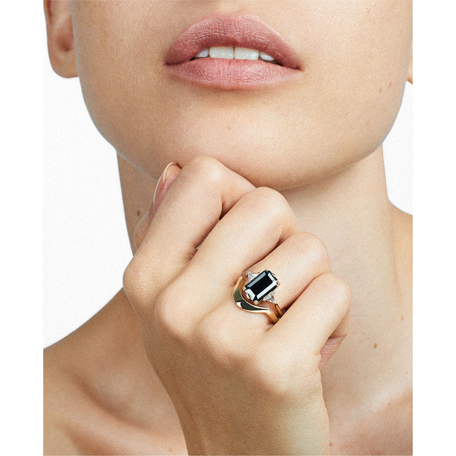 A three stone engagement ring with an emerald cut and emerald shape black diamond center stone with two white diamond trillion side stones set in yellow gold stacked above a tapered solid gold band on a light-complexion model's hand.