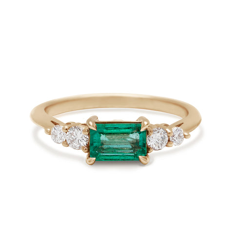 A ring with an emerald cut and shaped green emerald center stone with two round white diamonds on either side set in yellow gold.