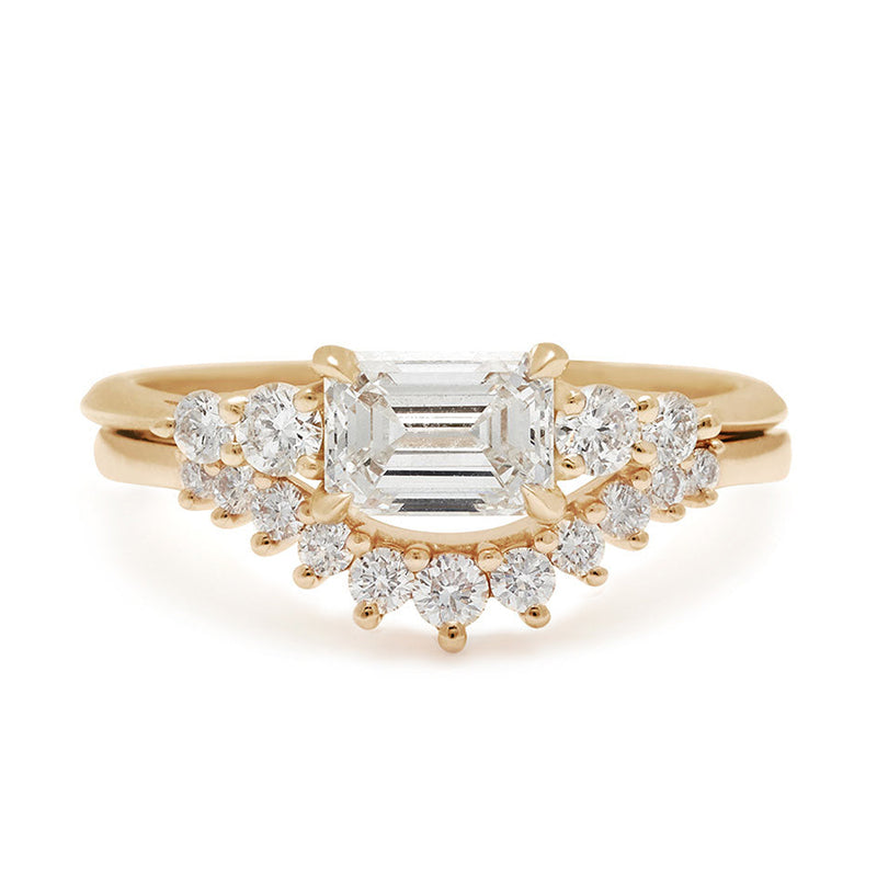 A ring with an emerald cut and shaped white diamond center stone with two round white diamonds on either side set in yellow gold set above an 11 white diamond curve band.