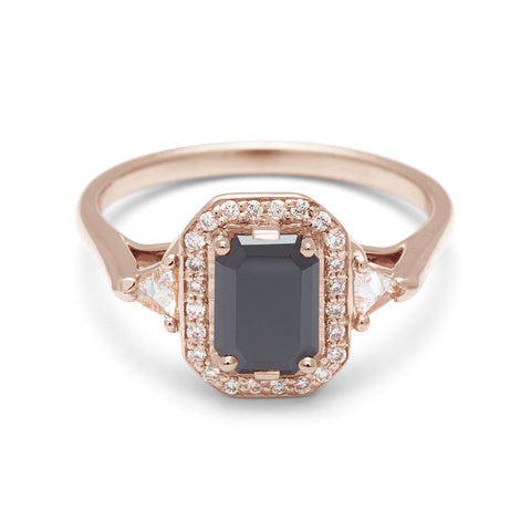 A three stone engagement ring with an emerald cut and emerald shape black diamond center stone surrounded by a white diamond pave halo with two white diamond trillion side stones set in rose gold., Rose Gold
