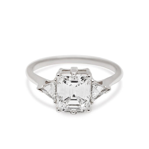 A three stone engagement ring with an emerald cut and emerald shape white diamond center stone with two white diamond trillion side stones set in white gold.