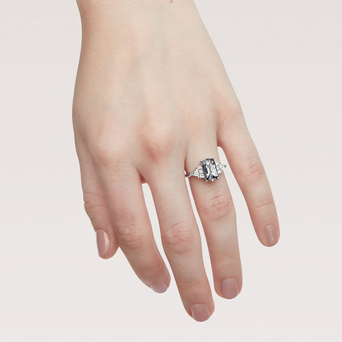 A emerald shape grey spinel center stone ring with three round white diamonds on either and two white diamond trillions on the outer edges of each side set in white gold on a light-complextion hand on a pink background. 3