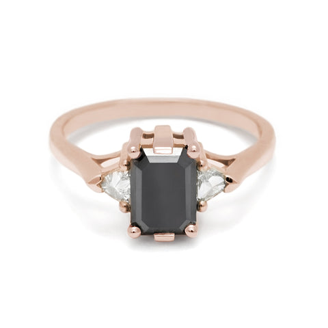 A three stone engagement ring with an emerald cut and emerald shape black diamond center stone with two white diamond trillion side stones set in rose gold.