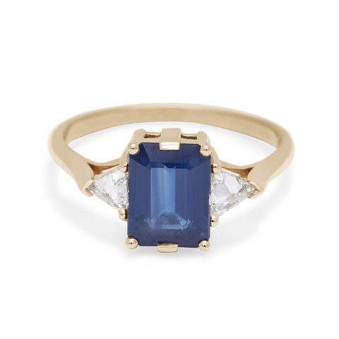 A three stone engagement ring with an emerald cut and emerald shape blue sapphire center stone with two white diamond trillion side stones set in yellow gold.