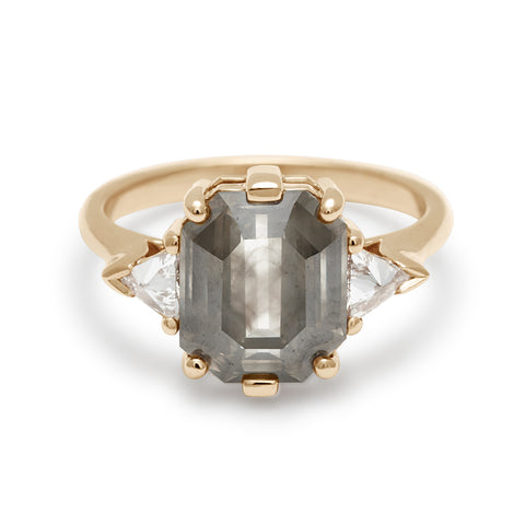 A three stone engagement ring with an emerald cut and emerald shape grey diamond center stone with two white diamond trillion side stones set in yellow gold.