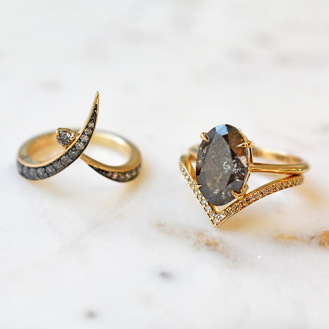 A solitaire ring oval grey diamond set in yellow gold set above a diamond dusted chrysalis shaped band and next to a wrap band on a marble stone.