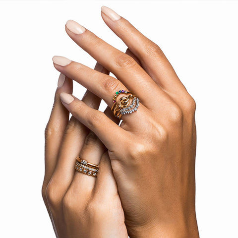 A curved 7 rainbow colored gemstones band set in yellow gold stacked above a serpent band and two white diamond curve bands on a medium-complexion left hand ring finger. On the right hand are three straight diamond dusted bands. 4