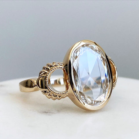 An oval rose cut bezel set white diamond set in yellow gold on a white table and grey background.