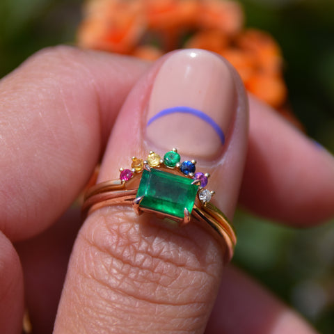 A curved 7 rainbow colored gemstones band set in yellow gold stacked above a east/west emerald shape green emerald ring on a light-compelxion thumb. 5
