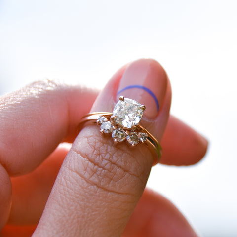 A curved 5 white diamond band set in yellow gold below a knife edge ring with a white diamond center stone on a light-complexion hand. 4