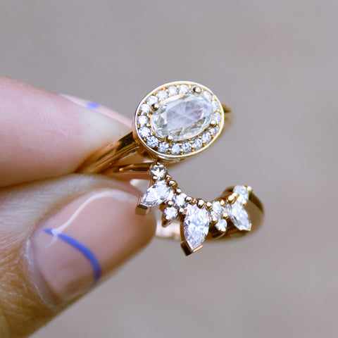 A hand holding a band with three marquise shaped white diamonds with round white diamond set in yellow gold and an oval halo engagement ring. 5