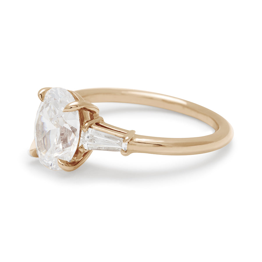 Angled view of a three stone engagement ring with an oval white diamond center stone and two white diamond tapered baguettes on either side set in 14k gold.