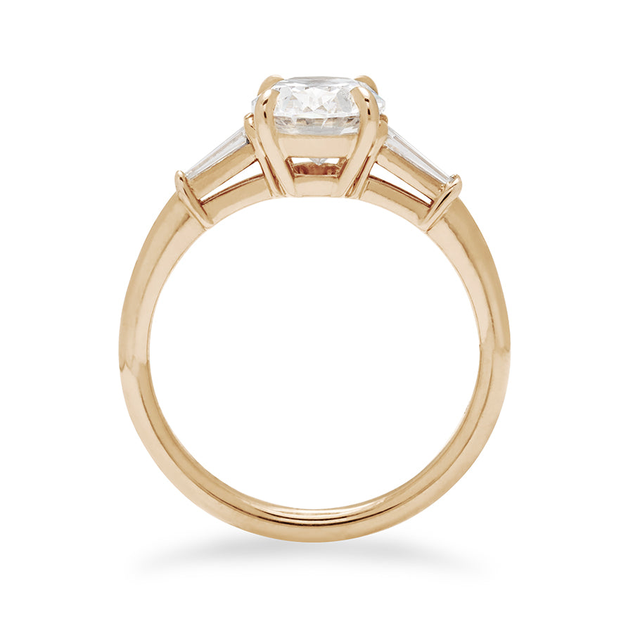 Side view of a three stone engagement ring with an oval white diamond center stone and two white diamond tapered baguettes on either side set in 14k gold.