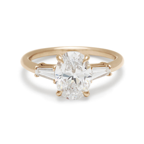 A three stone engagement ring with an oval white diamond center stone and two white diamond tapered baguettes on either side set in 14k gold.
