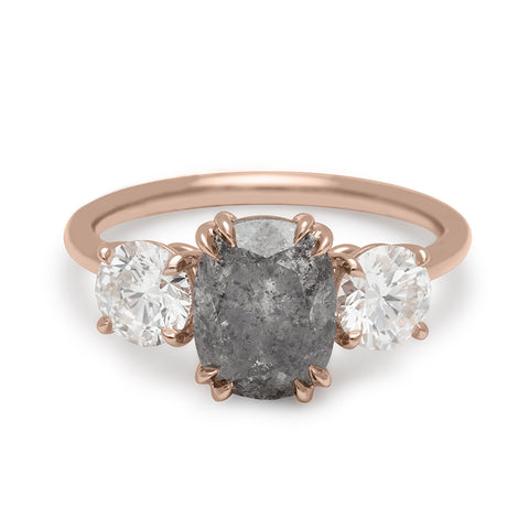 A three stone engagement ring with a cushion cut grey diamond center stone with two white diamond round side stones set in rose gold.