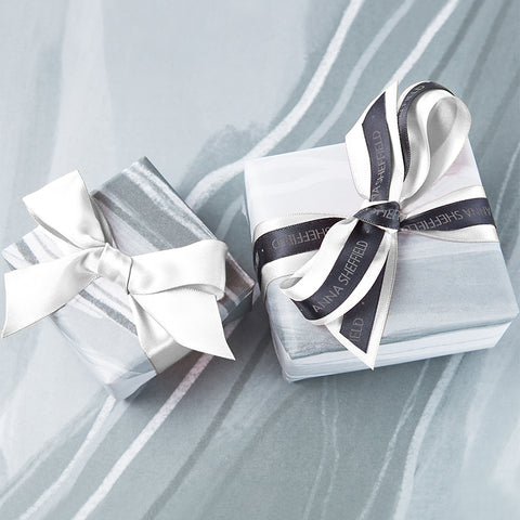Two boxes with grey and white gift wrapping and white bows one with "Anna Sheffield" on them on a blue grey background.