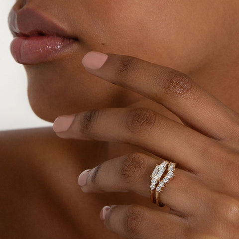 A ring with an emerald cut and shaped white diamond center stone with two round white diamonds on either side set in yellow gold above a 7 round white diamond curve band on a darker complexion model., White Gold, Rose Gold, Yellow Gold 2