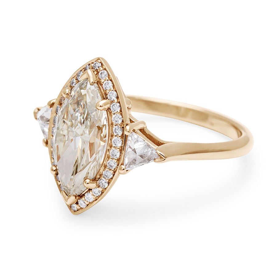 Angled view of a three stone engagement ring with a marquise shape champagne diamond center stone surrounded by a white diamond pave halo with two white diamond trillion side stones set in yellow gold.