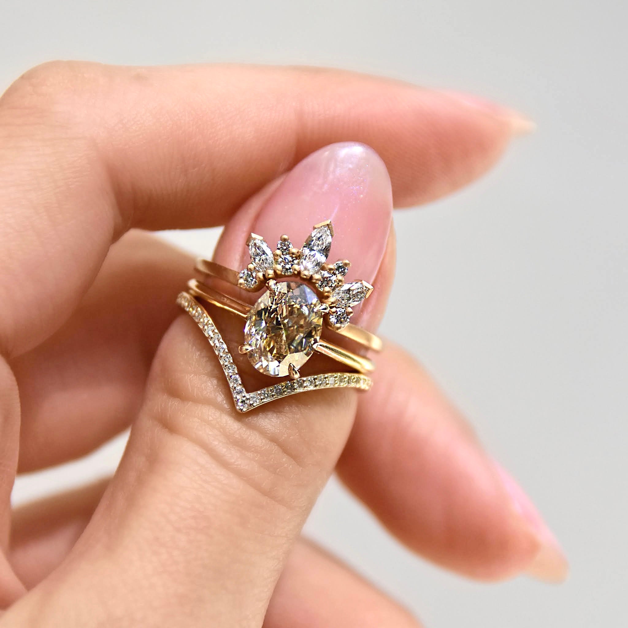 A band with three marquise shaped white diamonds with round white diamond set in yellow gold stacked above a solitaire oval champagne diamond engagement ring and a yellow gold nesting band on a light-complexion thumb., Yellow Gold, Rose Gold, White Gold