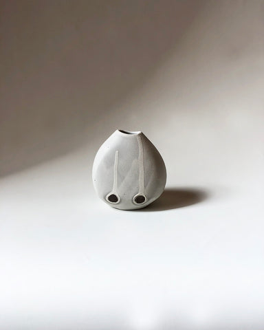 A ceramic pot in a blend of white and grey with two black dots at the bottom set on a grey background.