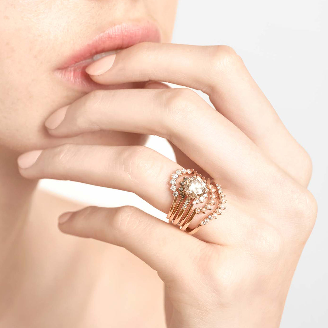 A curve band with 7 white diamonds set in yellow gold stacked above a smaller 7 diamond band, a solitaire champagne diamond engagement ring and three more curved diamond bands in assorted diamonds set in Yellow Gold, Rose Gold, and White Gold.